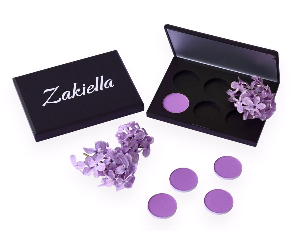 Empty magnetic eyeshadow palette shown closed with the Zakiella logo. Beside it is another magnetic palette with one purple eyeshadow placed and lilac flowers. There is also another bunch of lilac flowers below and more purple eyeshadow (called Celebration) pans. These palettes are for custom eyeshadow palette creations using our natural and organic clean formulas.